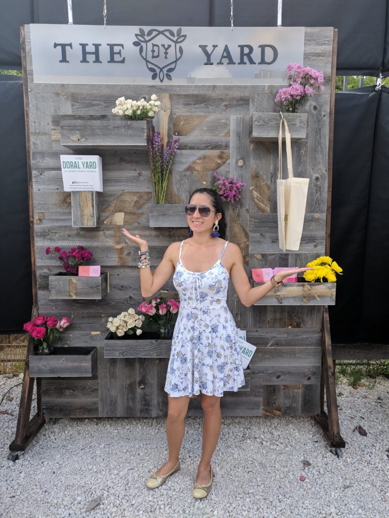Lilia Pozos stands in front of a wood board that showcases flowers and pamphlets. It reads "the doral yard" at the top. 