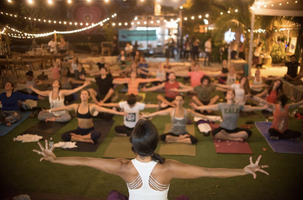 A lawn full of yogis, sitting cross legged with arms outstretched and eyes closed. The instructor's back is to the camera, also with outstretched arms. 