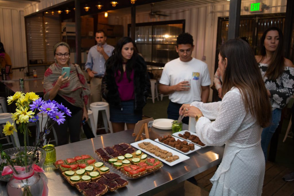 Lilia, wearing a white dress, stands over platters of sample foods. She uses tongs to fill small plates with food as the workshop attendees wait.