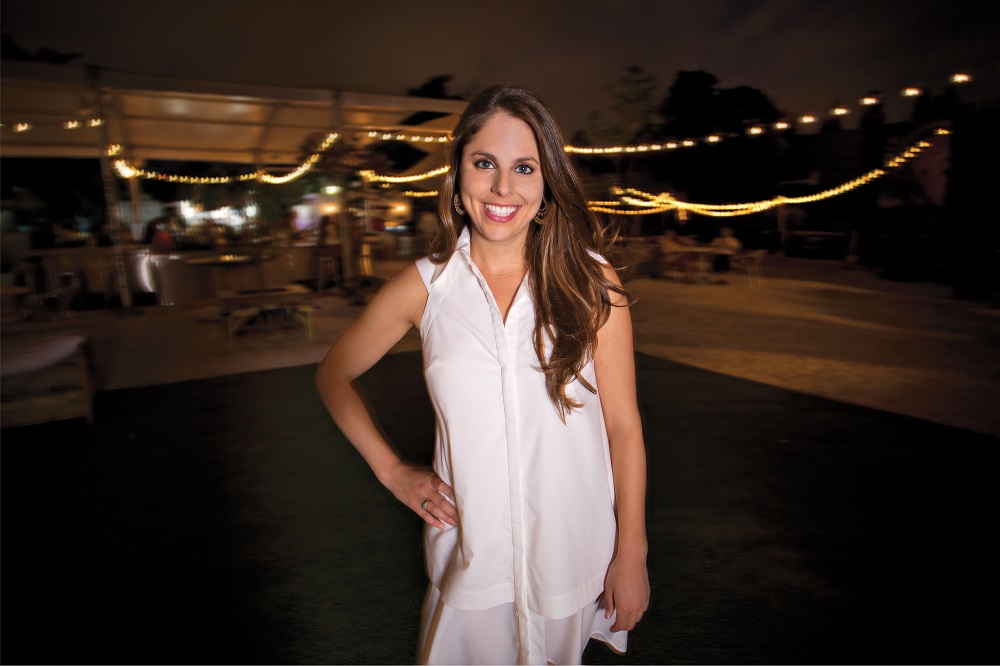Della Heiman stands in a white dress at The Wynwood Yard at night. String lights and a bar are visible in the background. 