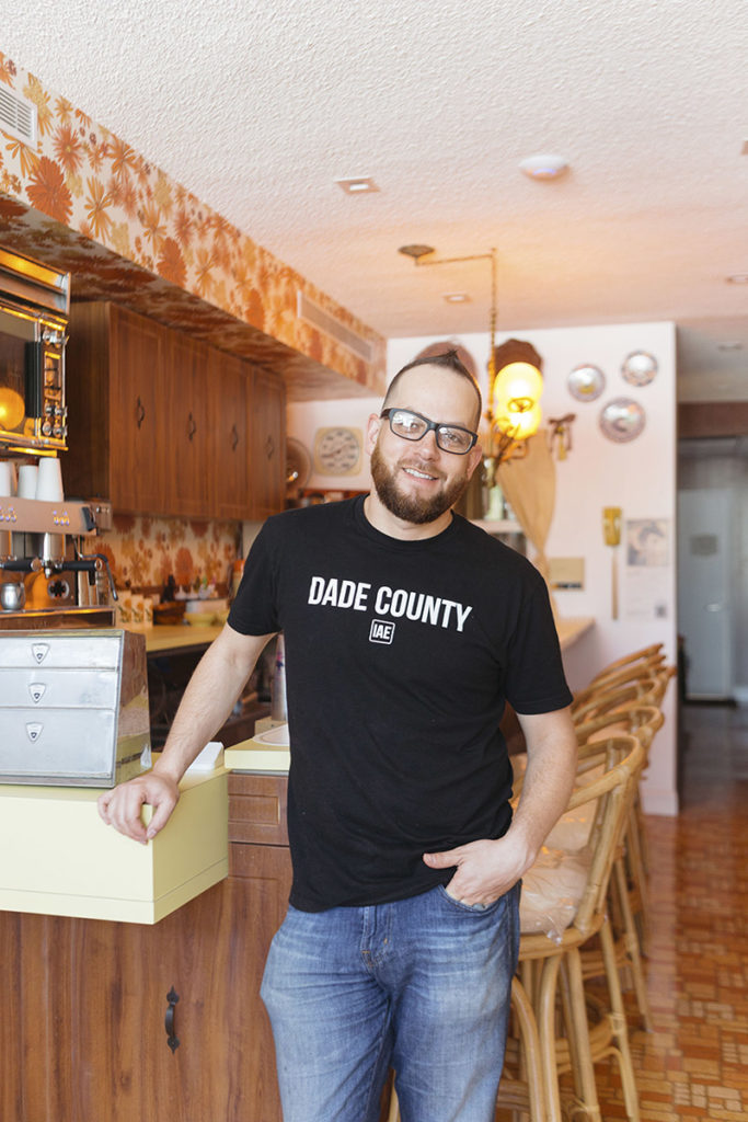 Matt Kuscher stands in a black t-shirt and jeans in front of a 1980's style kitchen. His restaurant, Vicky's House, is modeled after his childhood home. 