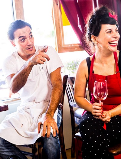 Benoit Rablat and Sandy Sanchez, the co-founders and partners behind La Fresa Francesa and Silverlake Bistro, sit together. Benoit wears an apron. Sandy wears a bright red apron and holds a wine glass. They're both  looking to the right and Sandy laughs.