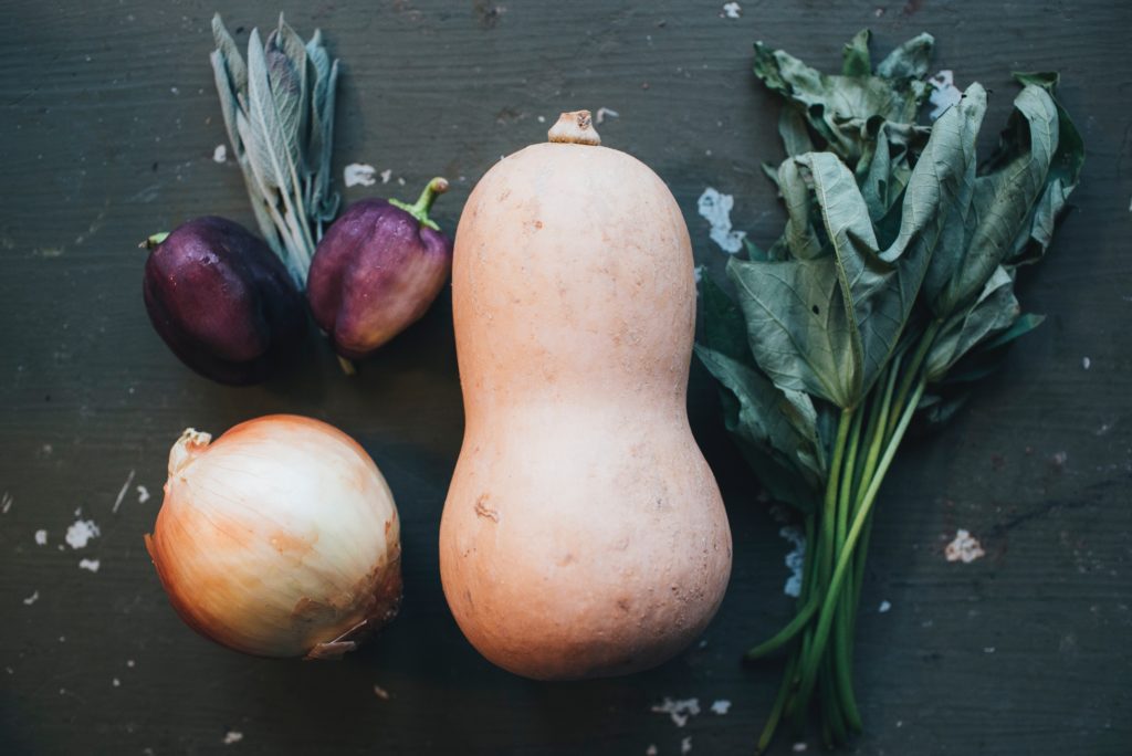 A butternut squash on a table, along with an onion, small eggplants, herbs and greens.