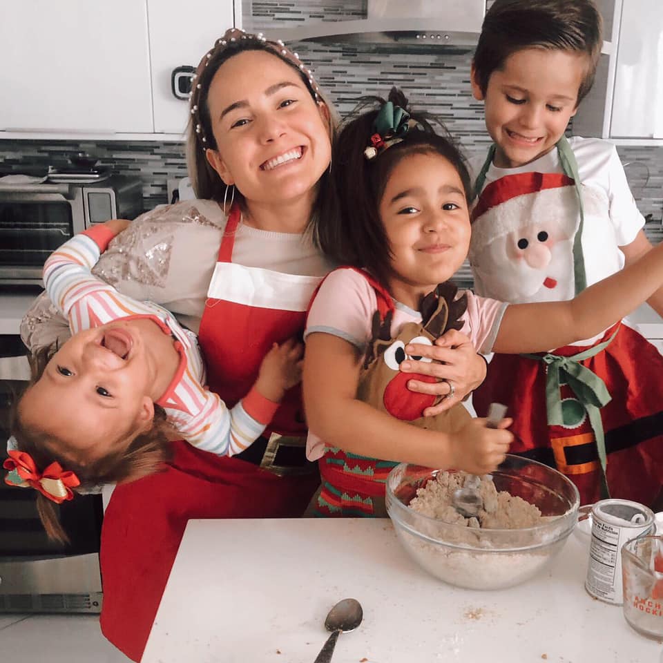 Fernanda Chacon, wearing a Santa apron, holds two girls, one of whom is stirring flour in a bowl. A boy stands next to them smilng. They also wear Santa aprons. Fernanda is the author of the article.