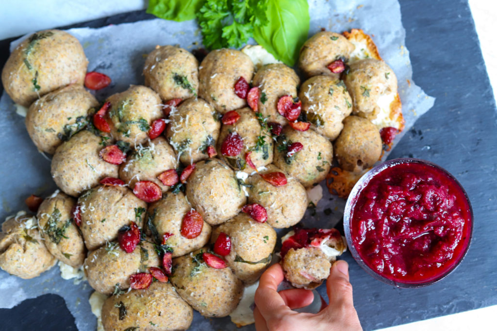 A hand grabs a piece of pull apart or monkey bread bread in the shape of a Christmas tree garnished with red cherries and green herbs on a granite slab. There is a small dish of cherry jam next to it. The image is to showcase the recipe offered here. 