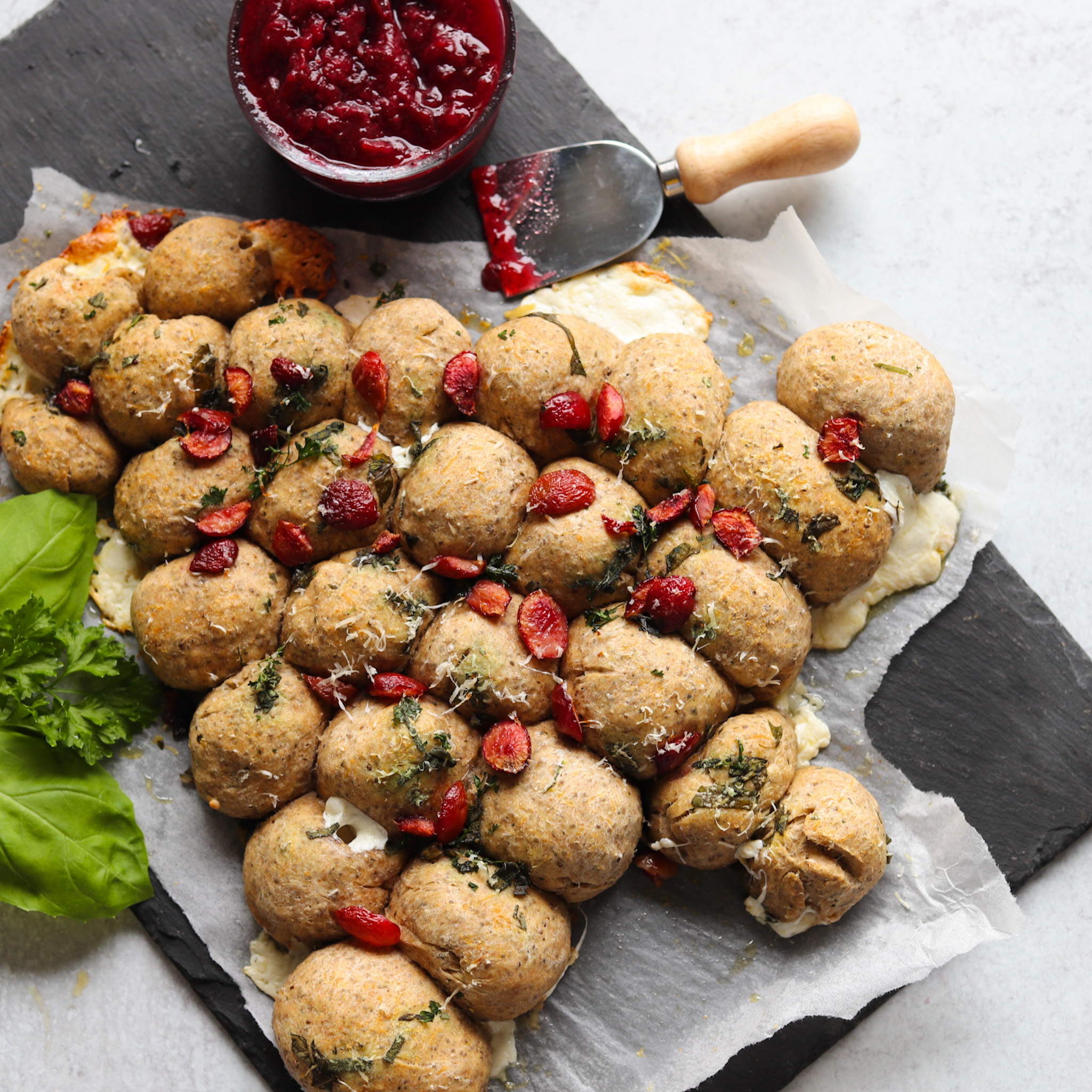 Pull-apart bread in the shape of a Christmas tree lays on wax paper on a granite slab. A serving knife with cherry jam lies next to it. This shows what the recipe for holiday pull-apart bread should look like when finished.
