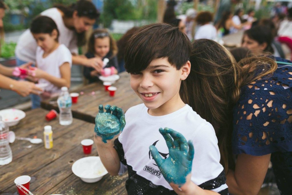 A smiling young boy holds his hands up covered in blue slime. 