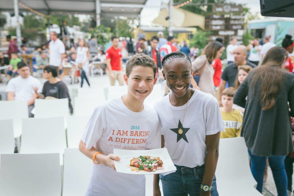 Two young entrepreneurs stand in front of an audience holding a plate of food.