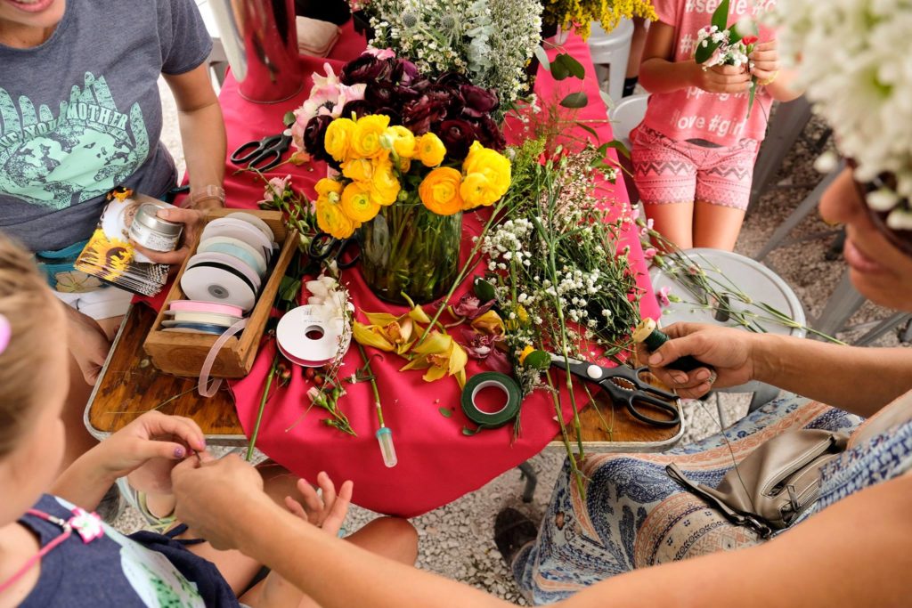 Cut flowers, ribbons, tools, and materials are on a table with active hands making flower crowns.