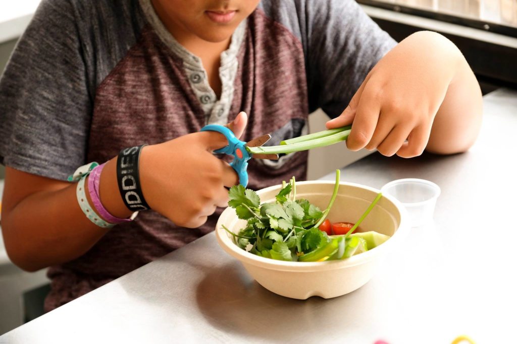 A boy uses scissors to make salsa in a bowl filled with fresh vegetables. 