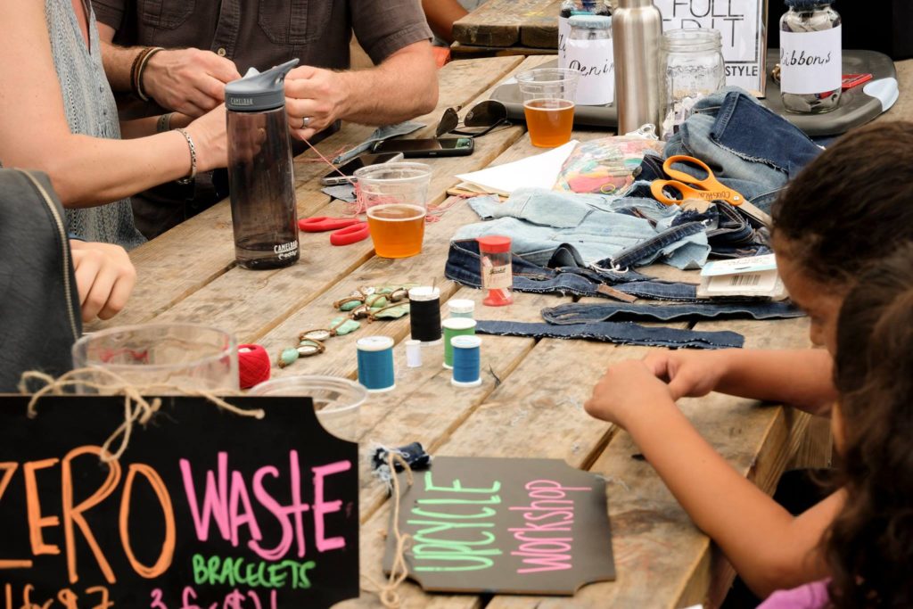 Kids and adults hands making denim bracelets on a wooden table. spools of thread, denim, and scissors are on the table. 