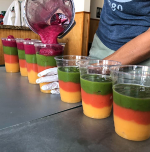 smoothies being poured into cups. The smoothies are layered in yellow, orange, green, and bright pink. 