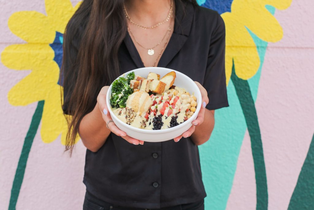 A della bowls team member holds a bowl layered with colors and textures in front of a mural